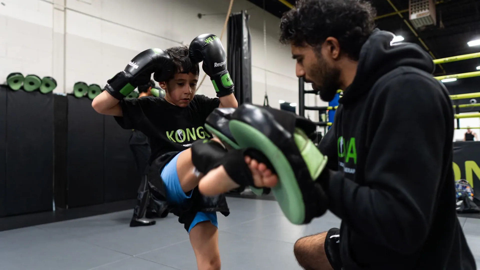 Child practising a knee strike against pads held by a coach