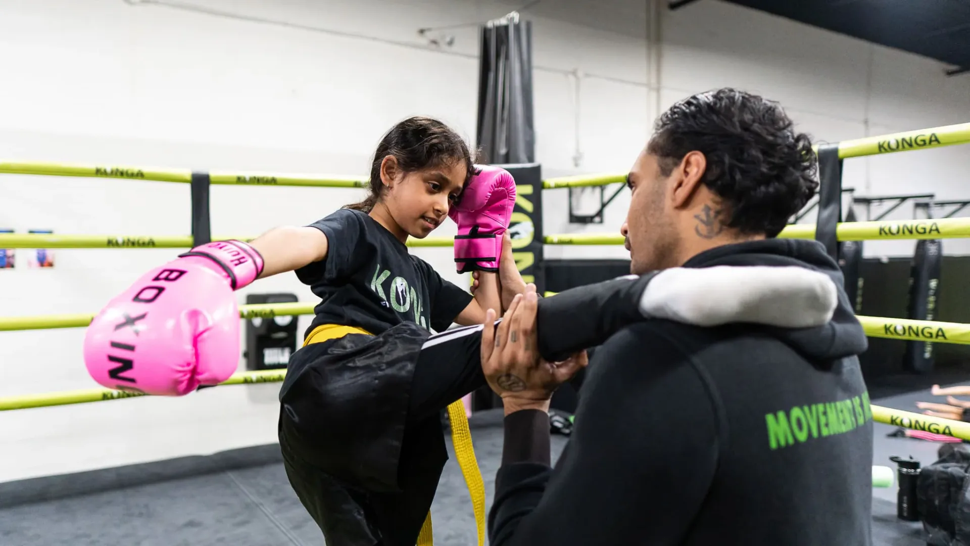 Child wearing pink boxing gloves practising a kick against a coach's pads