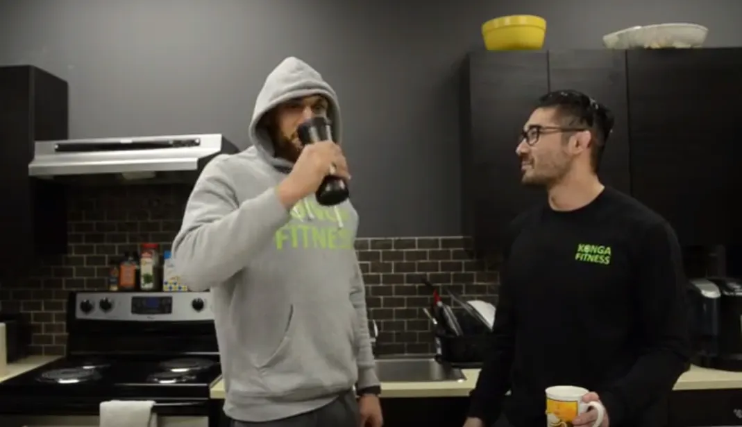 Two men in a kitchen, one drinking from a shaker bottle