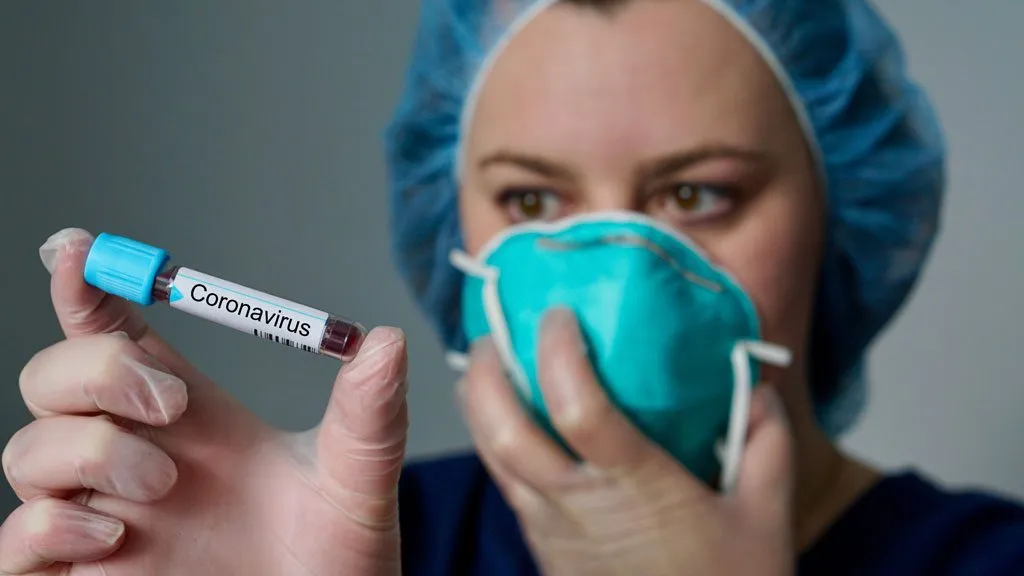 Person wearing a mask and holding a sample tube labelled Coronavirus